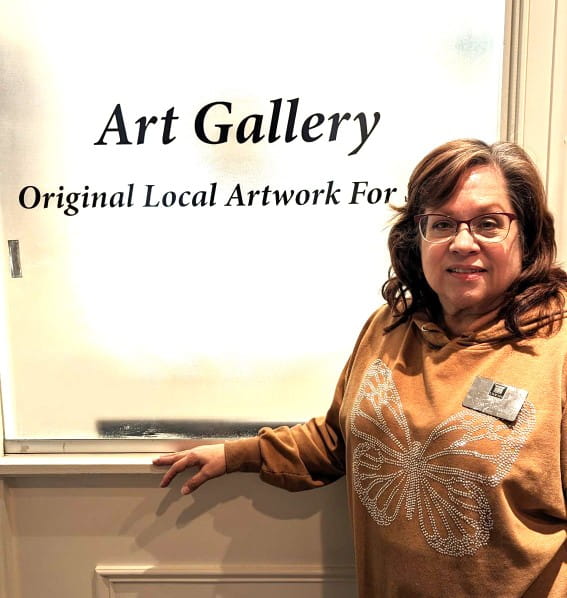 Cathy Eggleston the Gift Shop Manager standing in front of a board saying "Art Gallery / Original Local Artwork for Sale"